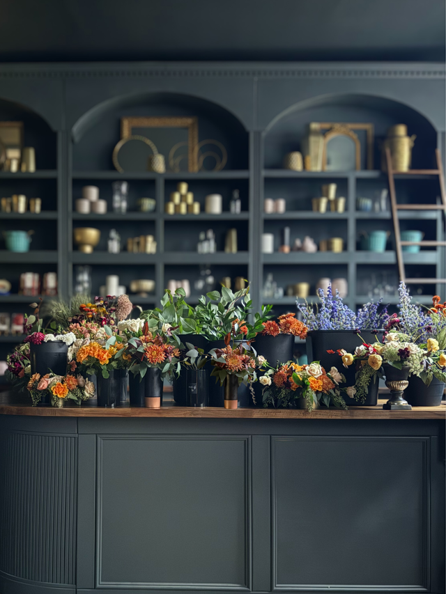 Floral arrangements on a counter with a dark green shelving unit in the background