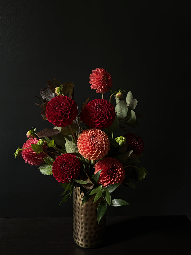 A bouquet of red and orange dahlia flowers with green foliage arranged in a hammered metallic gold vase against a dark black background.