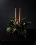 A dark green evergreen wreath centerpiece with two tall tan taper candles, small orange berries, and green leaves, set on a black surface against a black background.