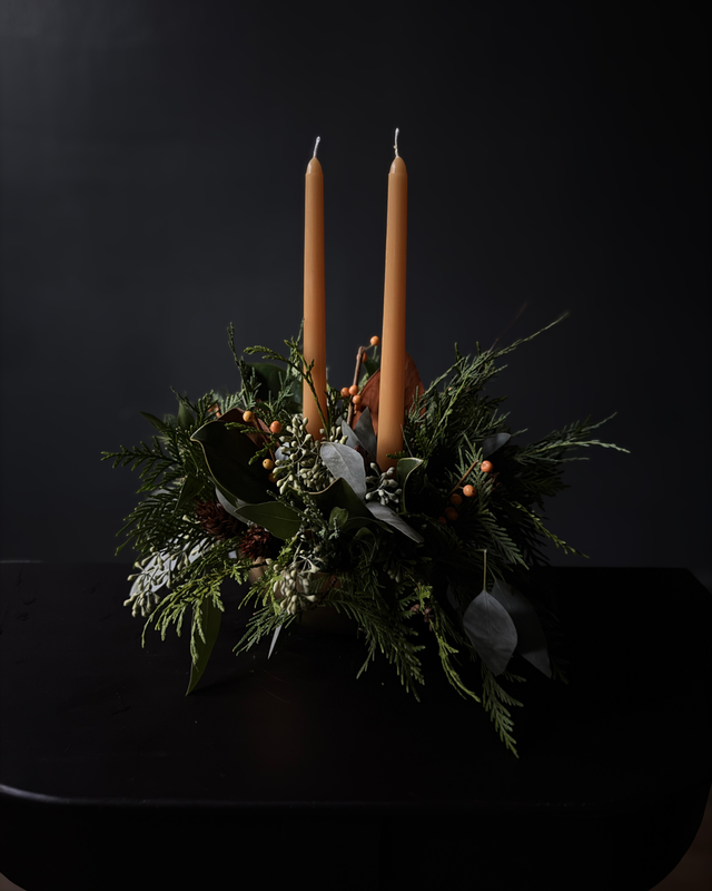 A dark green evergreen wreath centerpiece with two tall tan taper candles, small orange berries, and green leaves, set on a black surface against a black background.