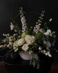 A large white and green floral arrangement with white roses, delphiniums, and hydrangeas in a matte white bowl vase against a dark background.