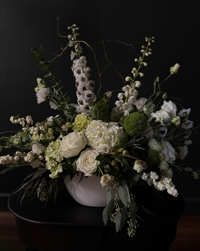 A large white and green floral arrangement with white roses, delphiniums, and hydrangeas in a matte white bowl vase against a dark background.