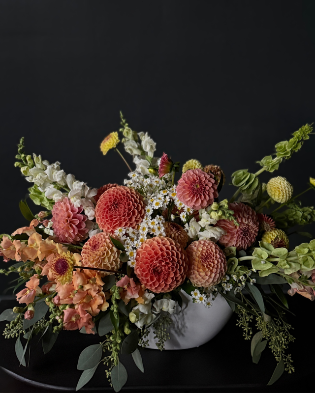 A rustic white ceramic vase filled with a dense, colorful autumnal floral arrangement featuring large coral dahlia, peach impatiens, white daisies, and green foliage