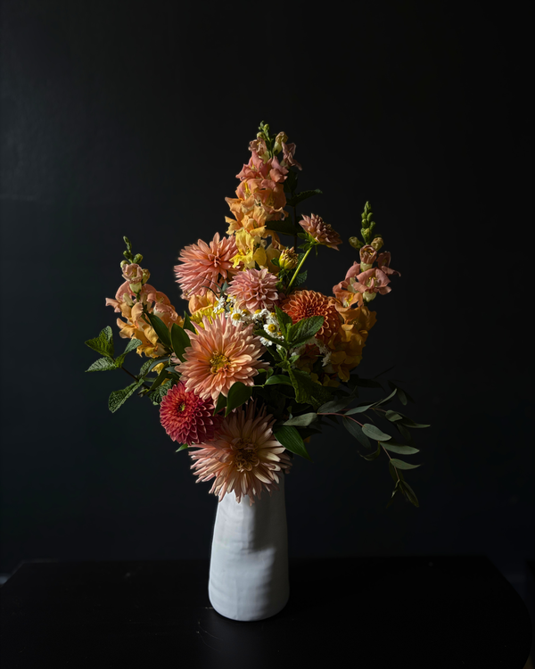 A floral arrangement featuring peach, orange, yellow, and red dahlia-like flowers with green foliage, presented in a small white ceramic vase against a dark black background.