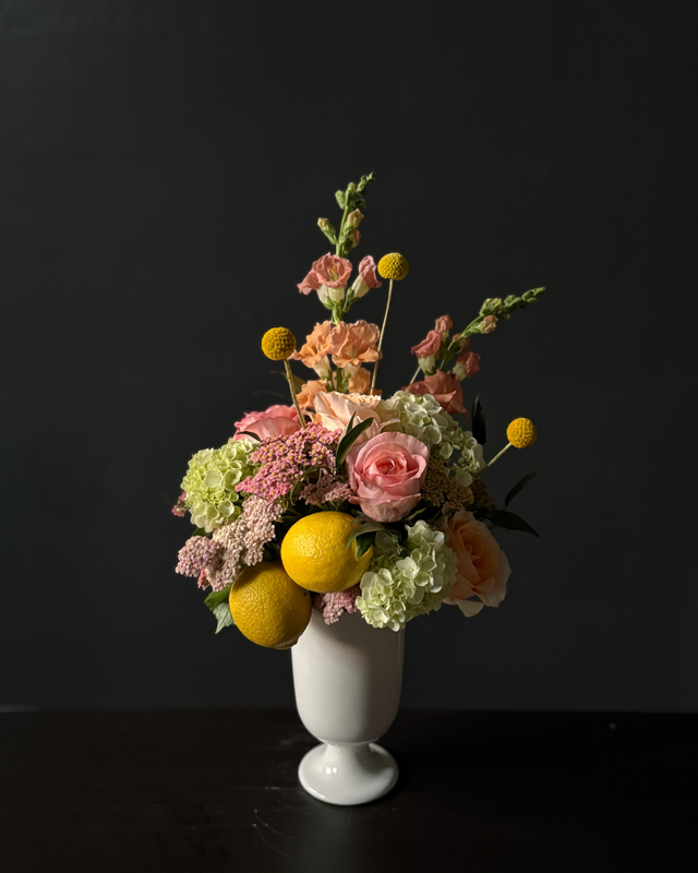 A floral arrangement in a white pedestal vase, featuring pink and peach roses, hydrangeas, snapdragons, yellow pom-pom flowers, and two lemons,