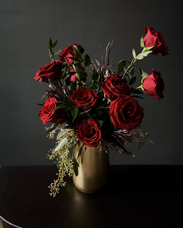 A bouquet of deep red roses with green leaves and dark filler stems arranged in a matte gold cylindrical vase,